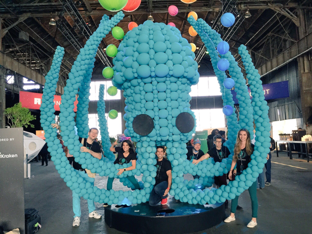 Six people pose around a large balloon sculpture of the GitKraken mascot, shaped like a squid with outstretched tentacles. The sculpture is displayed in a large indoor event space with colorful balloons suspended overhead.