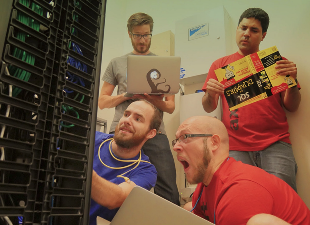 Four people crowd around a server rack in a small tech room. One person plugs in cables while another looks shocked. A third holds a laptop with a Kraken sticker, and the fourth reads a "SQL For Dummies" book.