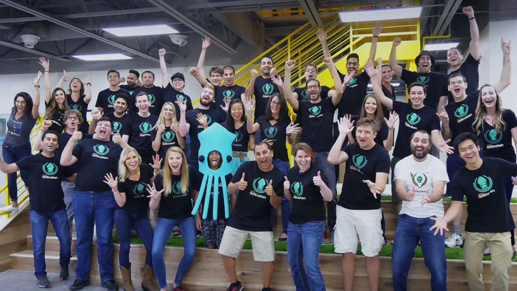 A large group of GitKraken team members pose on an indoor staircase, cheering and smiling, many wearing black GitKraken t-shirts. A person in the center wears a bright blue GitKraken squid mascot costume.